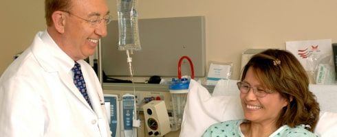 Smiling doctor in a lab coat conversing with a female patient in a hospital room with medical equipment in the background, reflecting compassionate healthcare.