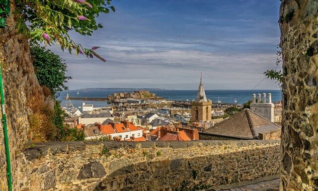 Picturesque view of St Peter Port in Guernsey, with the town's historical buildings, church spire, and harbour in the foreground, set against a backdrop of the English Channel and clear blue sky, framed by greenery and a stone wall.
