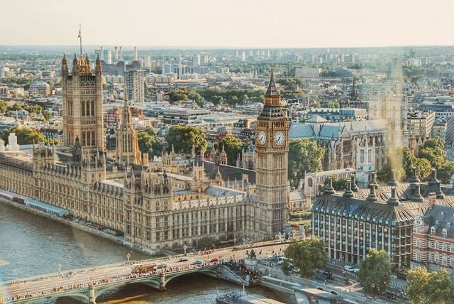Aerial view of the iconic Palace of Westminster and Elizabeth Tower, commonly known as Big Ben, by the River Thames in London, with a view of the cityscape and the Westminster Bridge teeming with pedestrians, on a sunny day.
