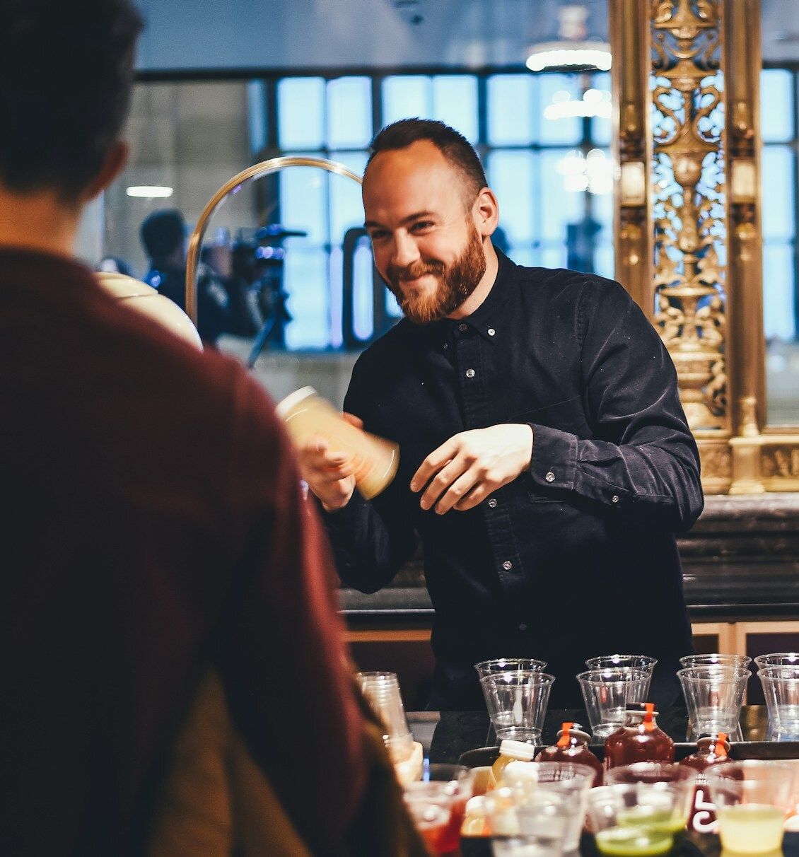 Smiling bartender engaging with a customer while expertly mixing a drink behind a bar adorned with a variety of colourful cocktail ingredients, reflecting skilled mixology and vibrant hospitality.