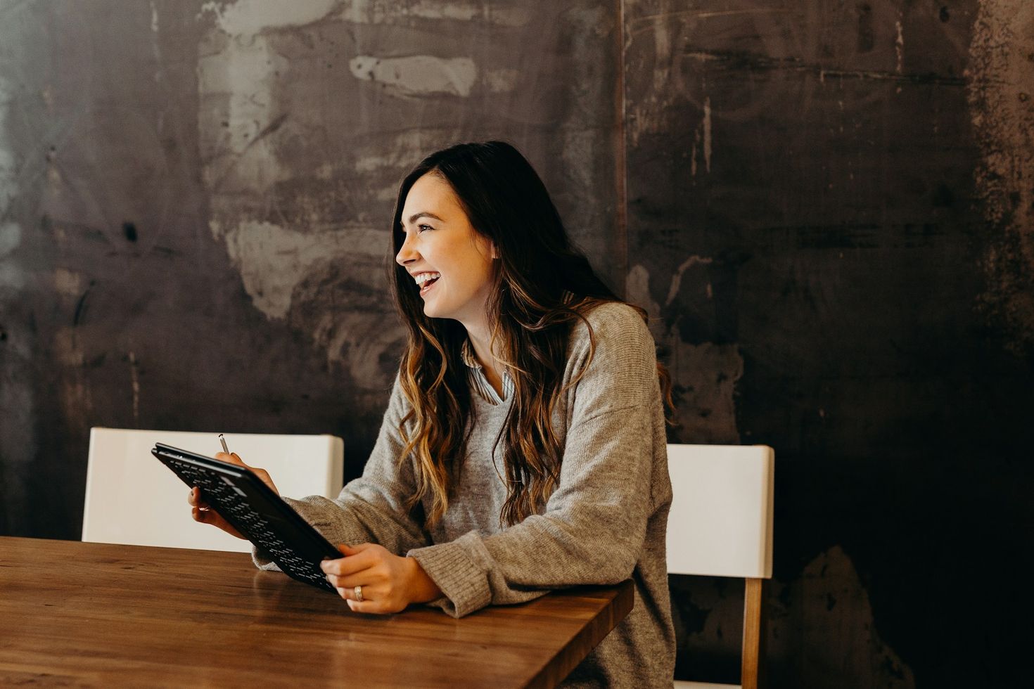 A joyful woman seated at a wooden table, holding a clipboard and engaged in conversation, in a stylish workspace with an artistic grey wall, embodying creativity and productivity.