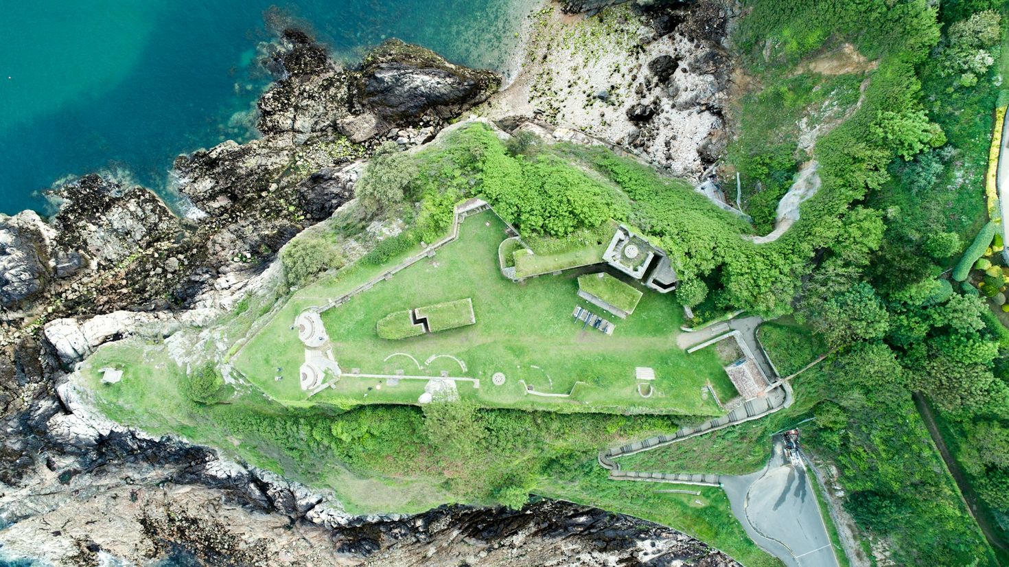 Aerial view of a historic Guernsey coastal fortification on a green headland, with rugged cliffs dropping into the turquoise sea, showcasing a blend of natural beauty and historical architecture.