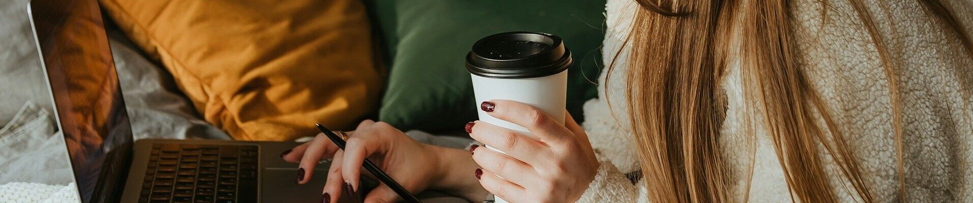 Close-up of a woman's hands with polished nails, holding a takeaway coffee cup and a pen over a laptop keyboard, indicating a comfortable remote work setting with warm tones.