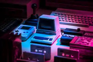 How to get started securing your business Retro tech-themed composition featuring vintage computers, a game console, joystick, and cassette tapes, all bathed in neon pink and blue lights, evoking nostalgia for the early era of personal computing and gaming.