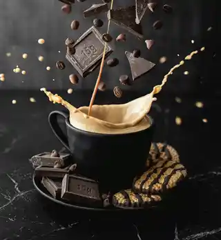 Dynamic image of coffee spilling over from a black cup with chocolate pieces and coffee beans suspended in mid-air against a dark background, creating a sense of movement and a delightful mix of flavours.