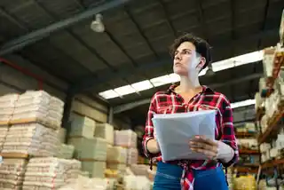 Focused manager holding papers in a warehouse, amidst stacks of inventory.