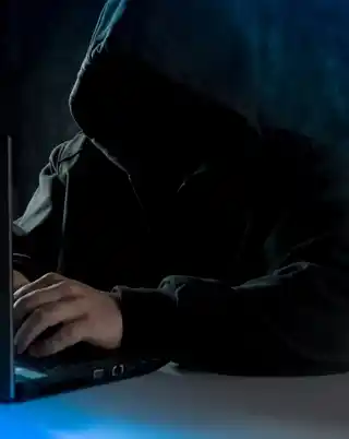 A hooded figure types on a laptop in a dark room, evoking themes of cyber security and online anonymity.