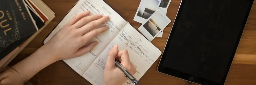Person writing in a diary with a tablet and instant photos on a wooden desk, planning and organising daily tasks and memories.