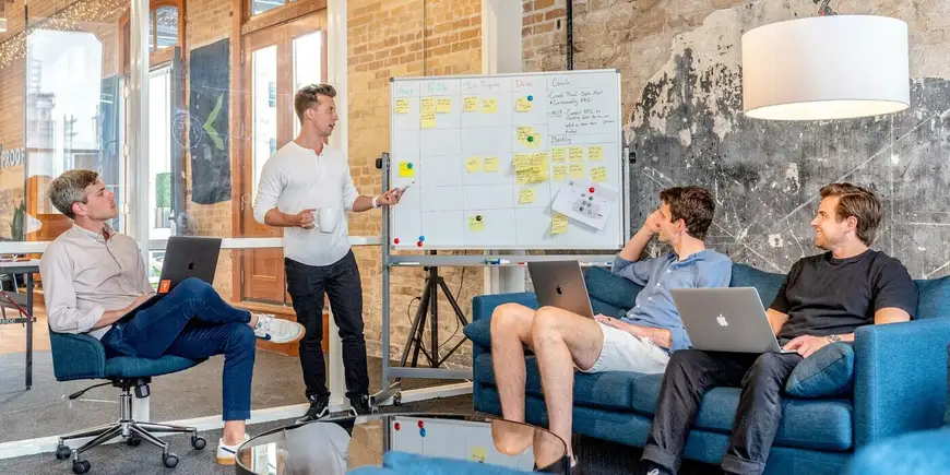 Business team in a strategy session in a modern office, with a presenter discussing project stages—Story, To Do, In Progress, Done, Goals—on a whiteboard, while colleagues collaborate on laptops.