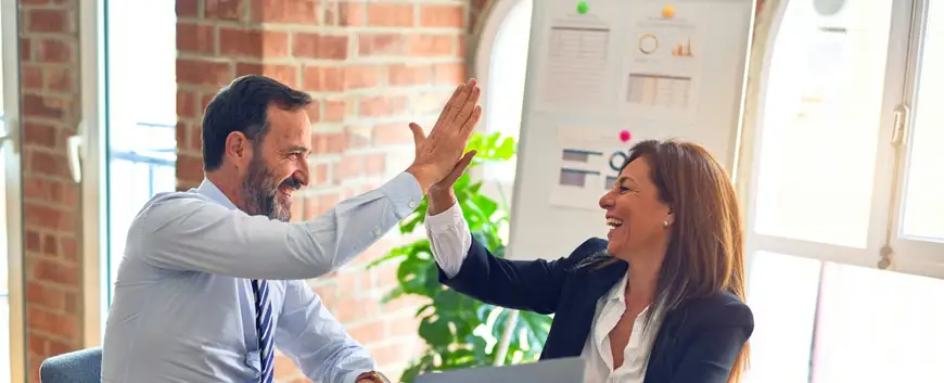 Two cheerful business colleagues giving a high-five in a bright office, celebrating success with visible joy, amidst a professional setting with brick walls and large windows.
