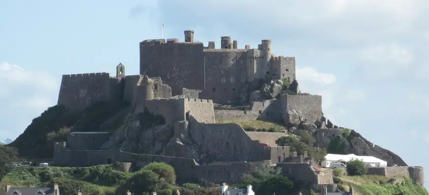 Daytime view of Mont Orgueil Castle in Jersey, dominating the skyline with its imposing medieval architecture on a hilltop overlooking houses and the sea, under a clear sky.